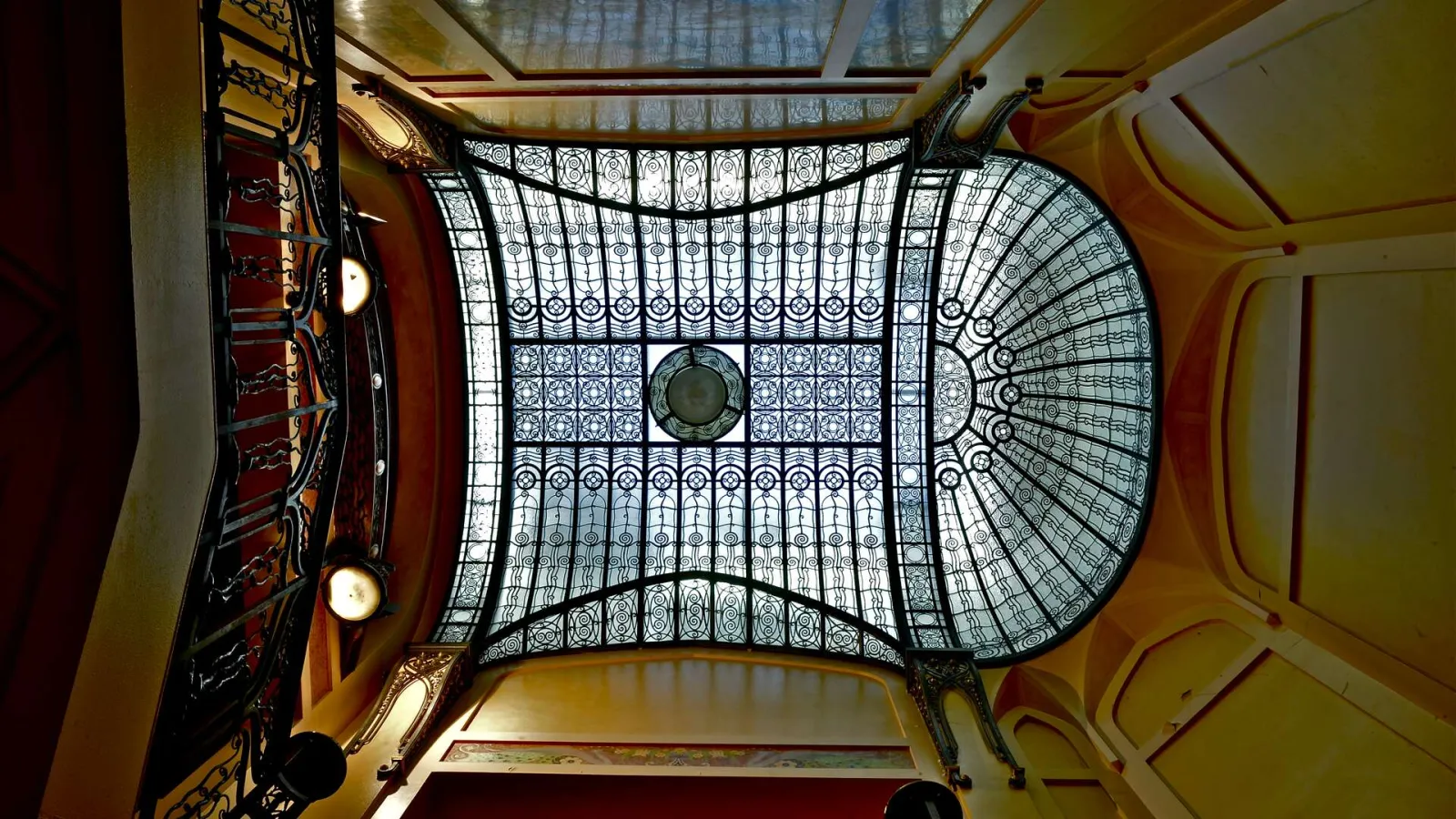 Overhead view of the stained glass ceiling at Hôtel Frugès in Bordeaux, displaying an ornate geometric structure with decorative patterns, gilded wooden frame, and Art Deco lighting fixtures.
