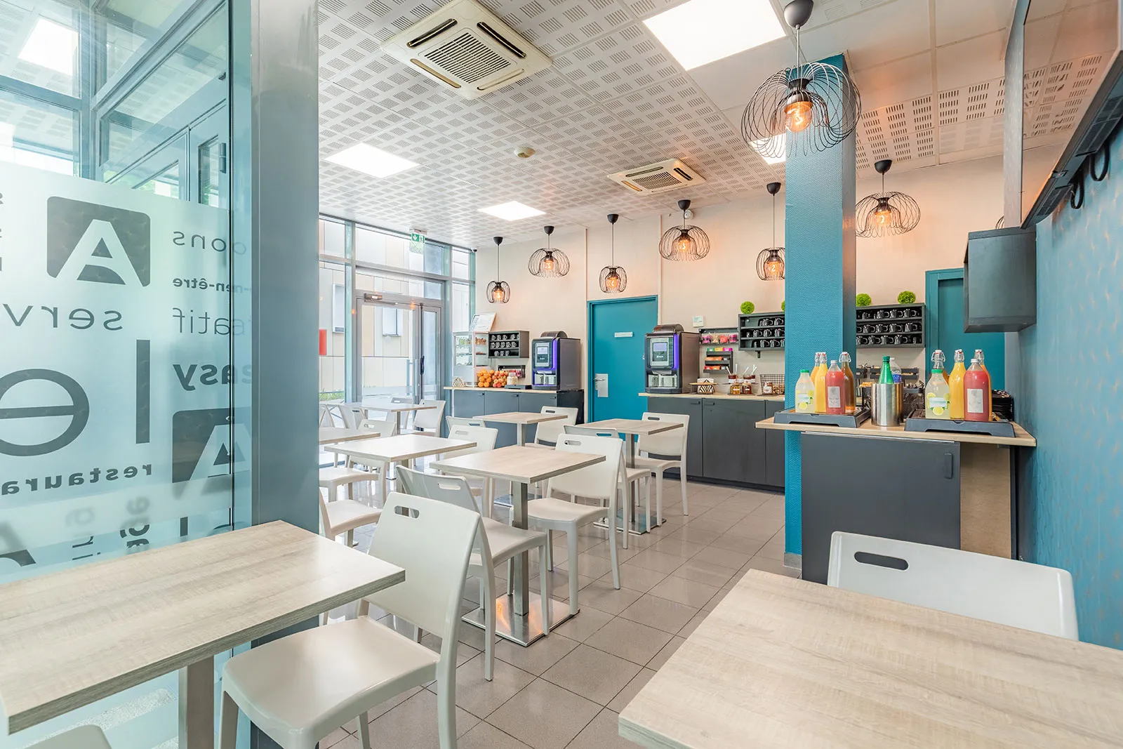 Modern common area of a serviced apartment in Bordeaux featuring an open kitchen, dining tables, white chairs, turquoise and grey furnishings, designer lighting, and large windows overlooking the street.