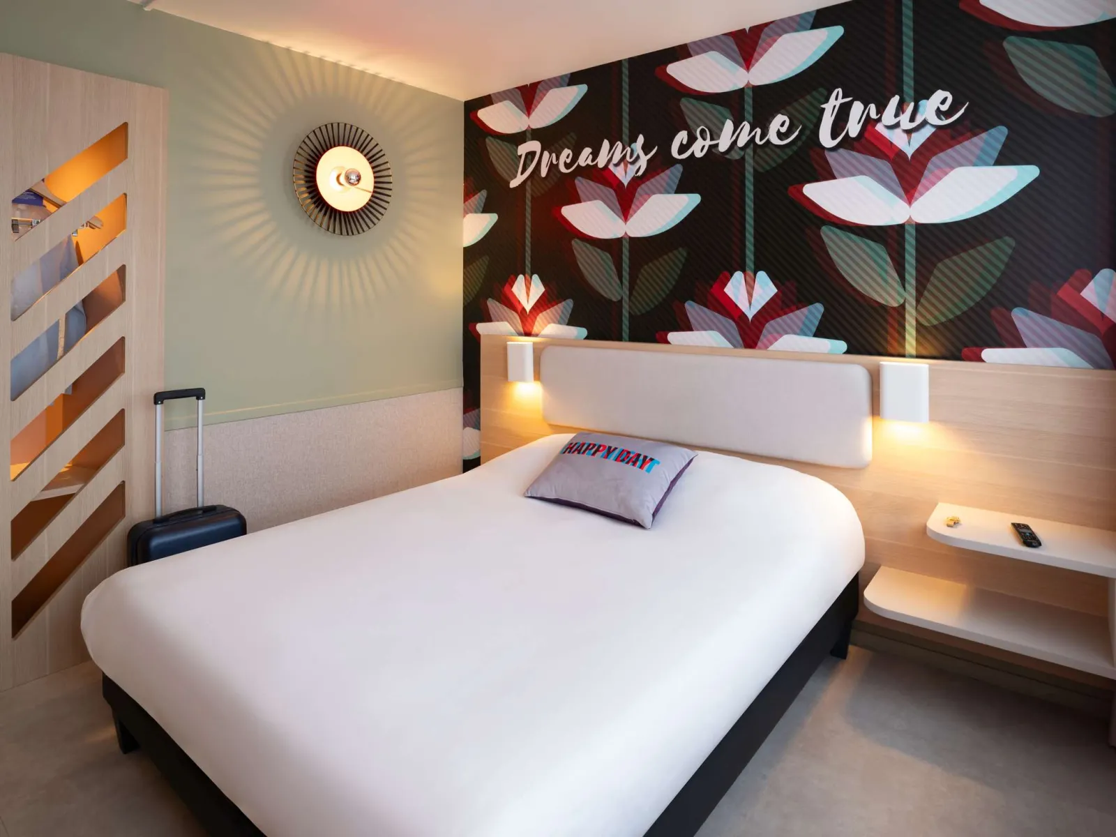 Modern hotel room featuring a white double bed, illuminated headboard, accent wall with red and white floral patterns, circular golden wall decoration, and inspirational text « Dreams come true »