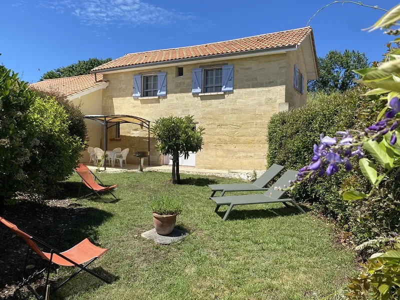 Rear view of a beige stone house with tiled roof, surrounded by a landscaped garden featuring lounge chairs, pergola, and lush vegetation, under clear blue sky.