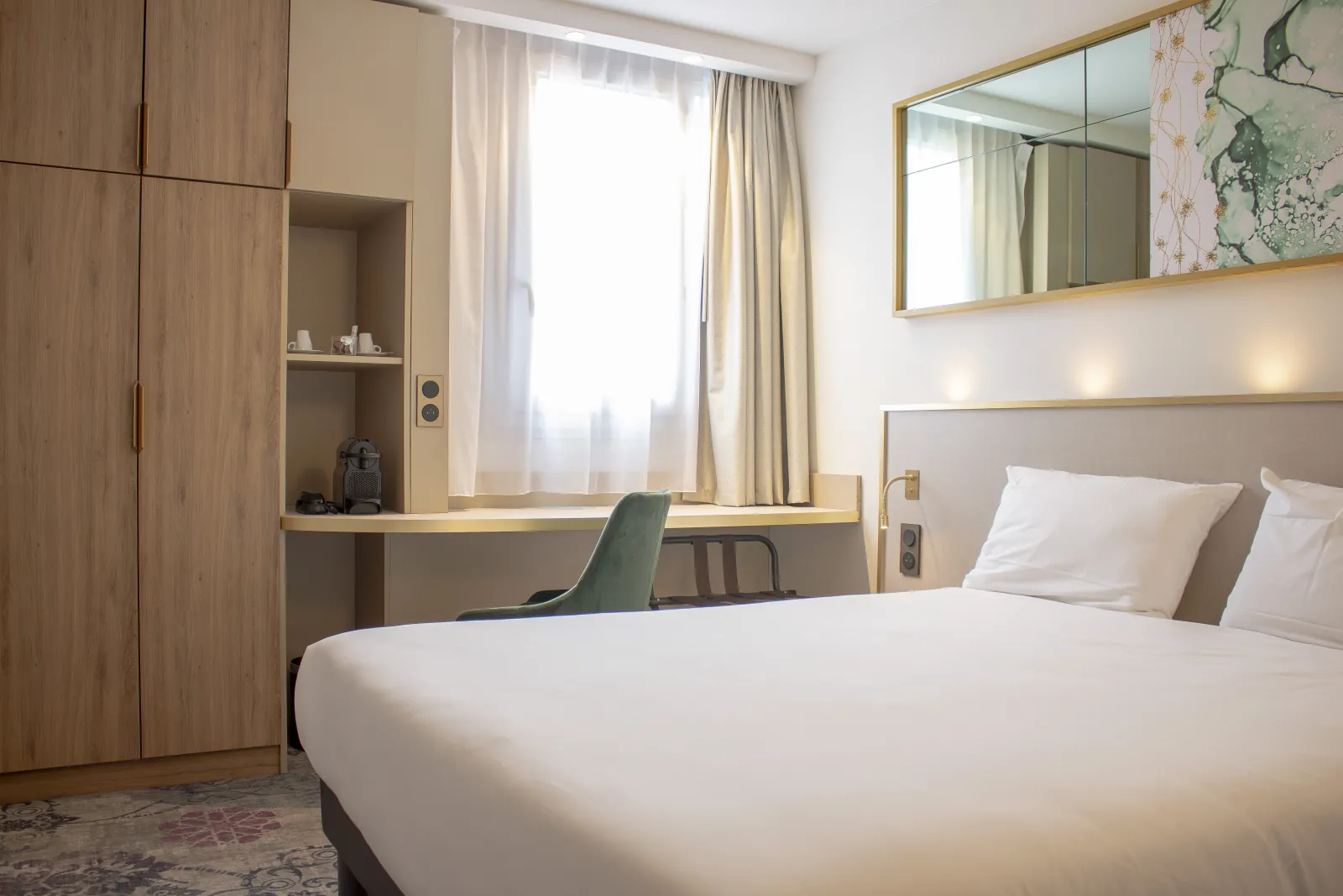 Modern hotel room featuring a double bed with white linens, integrated desk, light wood wardrobe, gold-framed mirror and window with beige curtains in Bordeaux