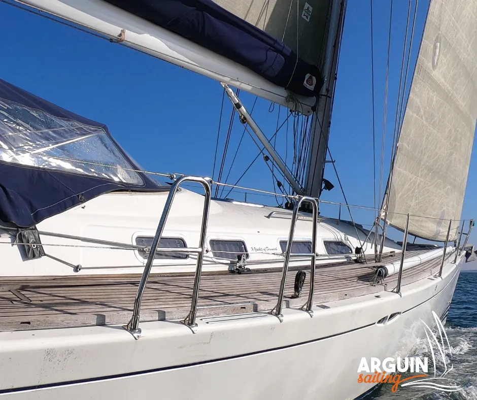 White sailing yacht with sails raised against clear blue sky, showing deck details and rigging, moored at Bordeaux with Arguin Sailing branding.