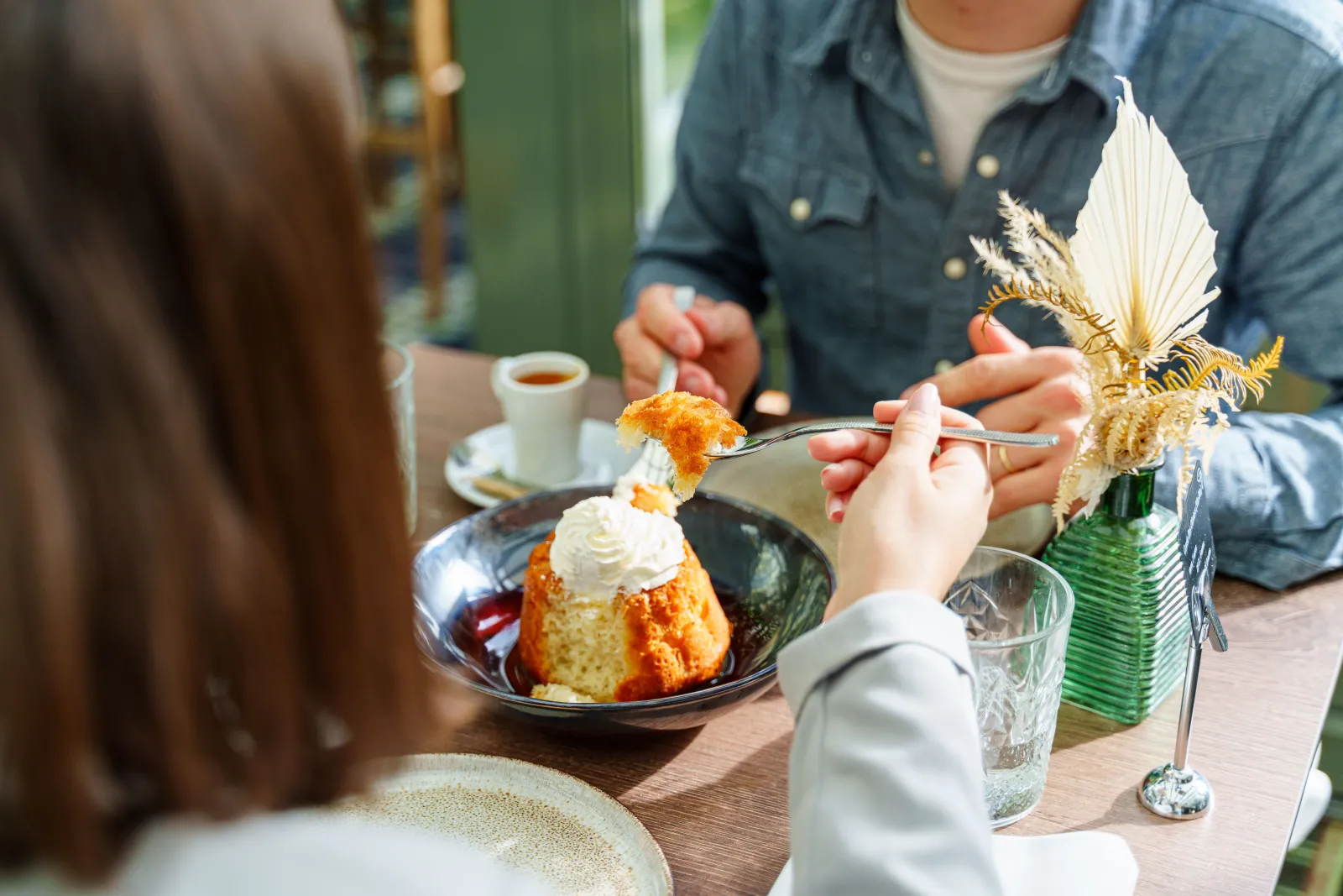 Two people enjoy a dessert at a restaurant. In the foreground, a woman uses a fork to take a bite of a cake topped with whipped cream and caramel sauce, served on a black plate. A white and gold flower garnishes the dish. In the background, a man in a blu