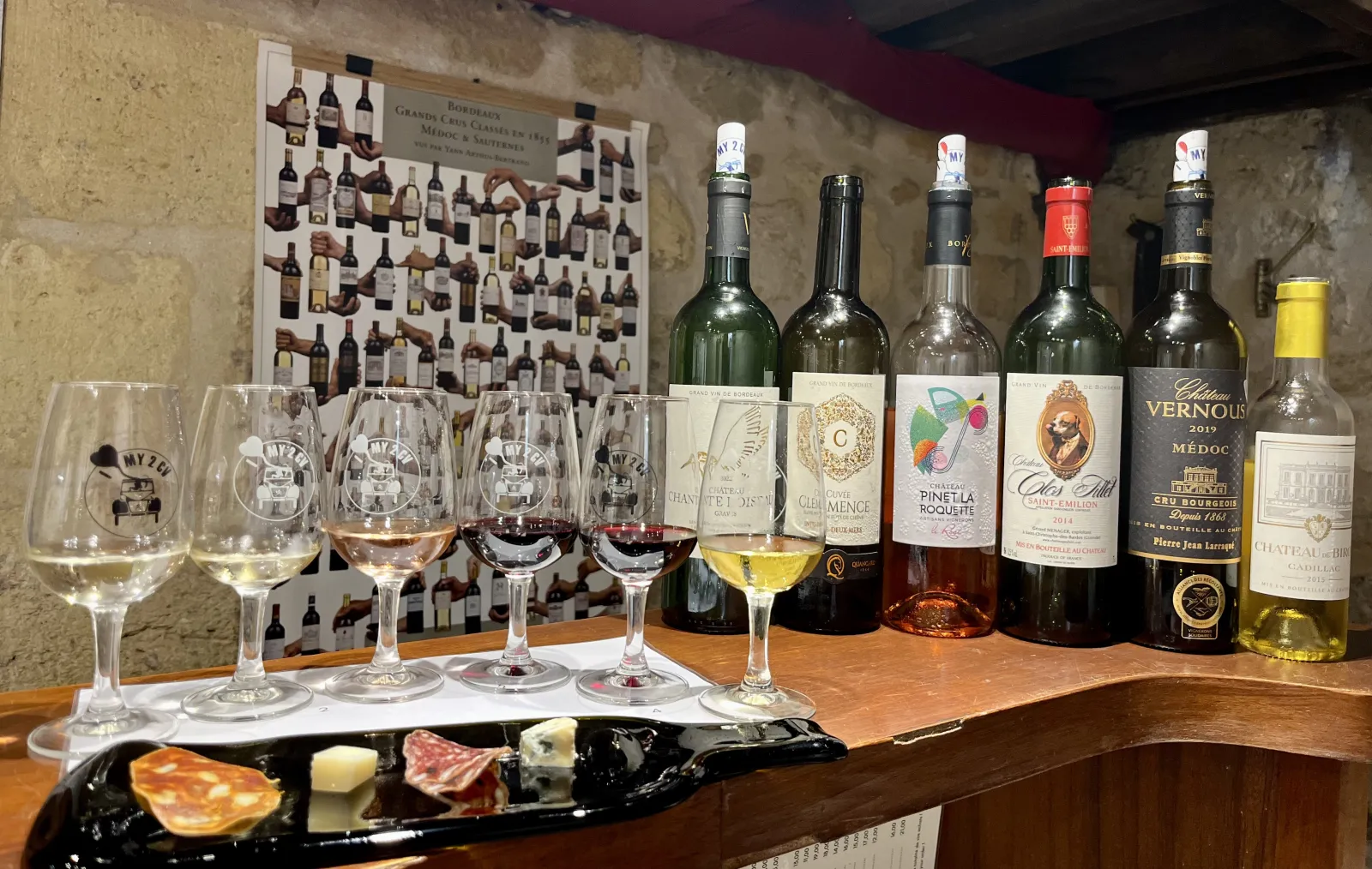 Bordeaux wine tasting featuring six glasses with different wines, six bottles displayed, a charcuterie and cheese plate, and a poster of Bordeaux Grand Cru Classés in the background.
