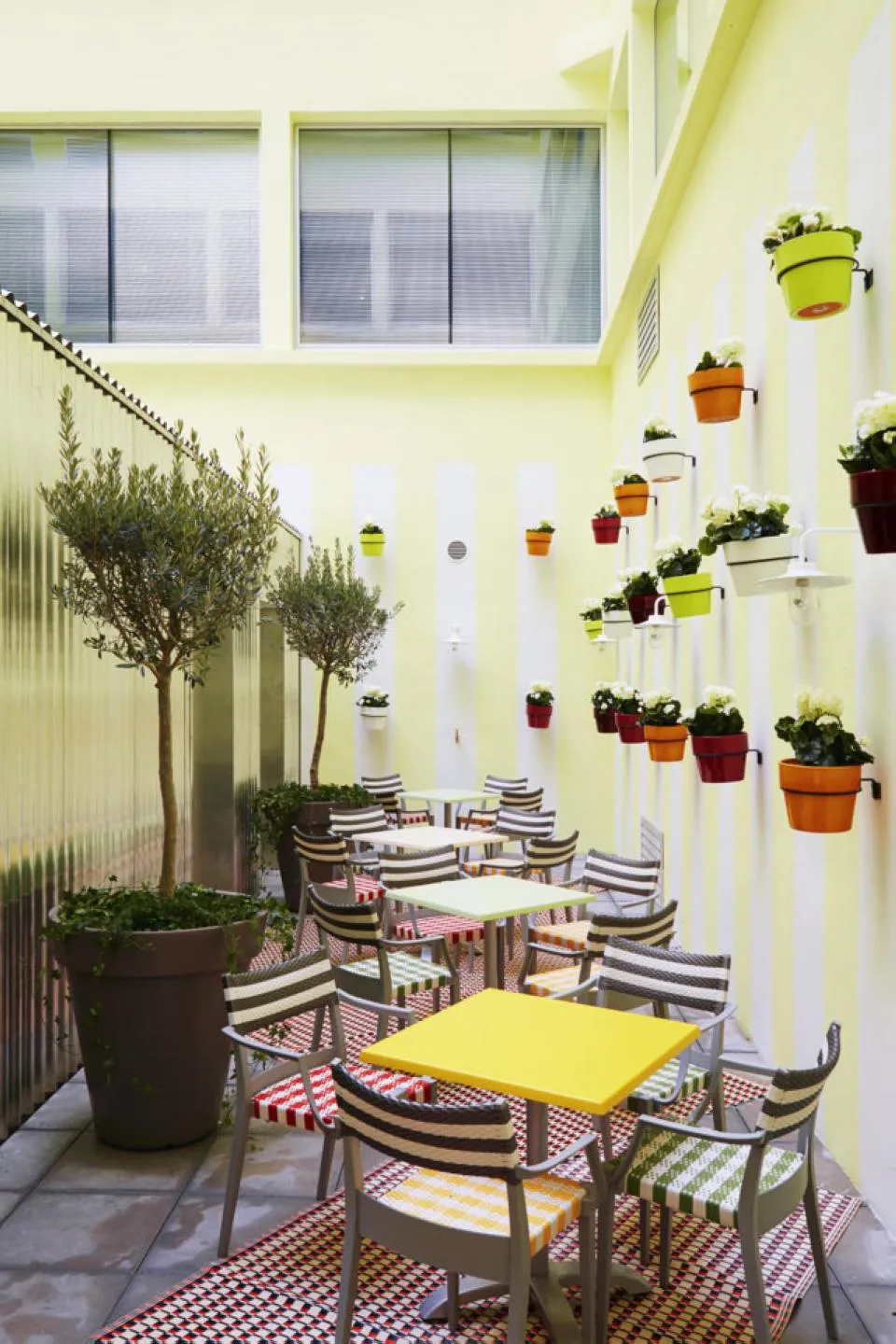 Bright interior terrace of Mama Shelter Bordeaux featuring pale yellow walls, striped blue and white chairs, yellow tables, hanging colorful flower pots, and potted trees.
