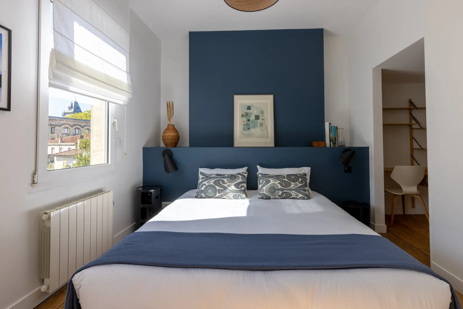 Modern bedroom in Bordeaux featuring a navy blue accent wall, double bed with white sheets and blue blanket, geometric patterned pillows, window overlooking Bordeaux architecture, white radiator, wall shelf with art objects, and open doorway to dressing r