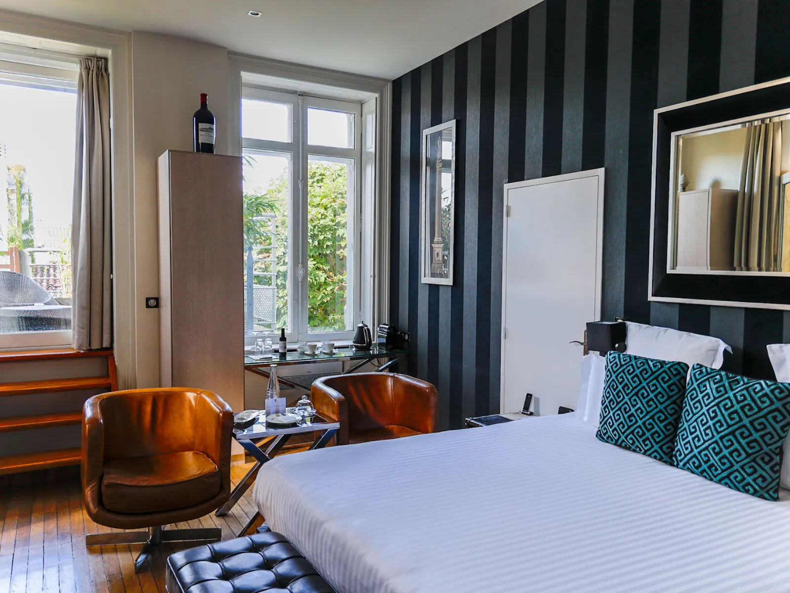 Elegant hotel room featuring a king-size bed, cognac leather chairs, black and grey striped accent wall, panoramic windows overlooking a terrace, and contemporary upscale decor