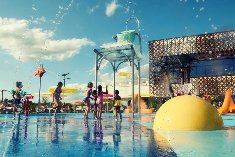 Children playing in a water park with water sprays, yellow dome and play structures under a sunny blue sky