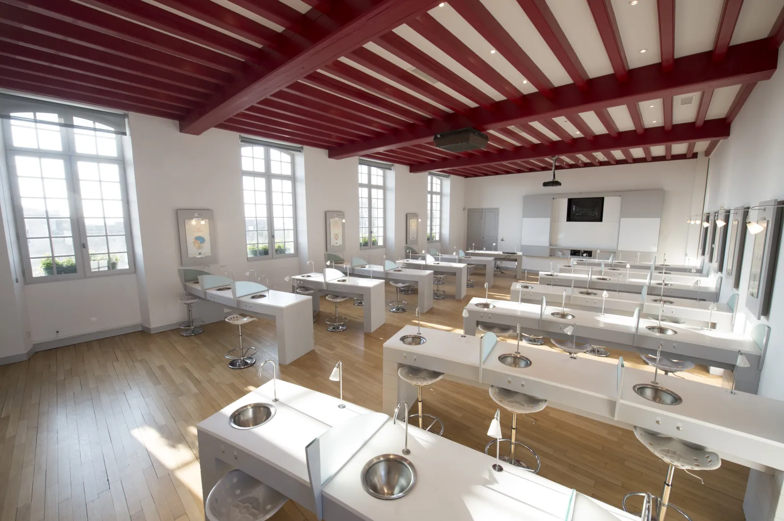 Modern interior of a wine tasting room featuring white workstations with wine glasses, red and white beamed ceiling, large windows, mirrors and chrome stools at the Maison du Vin de Saint-Emilion in Bordeaux.