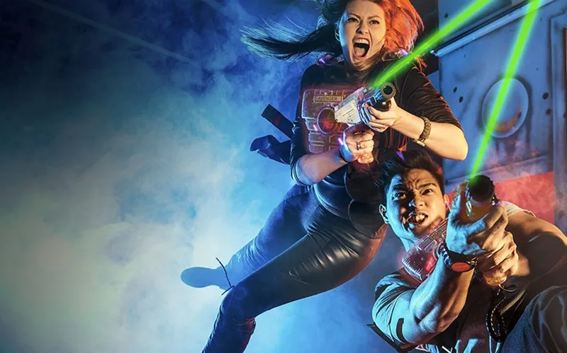 Three children playing with laser guns in a virtual reality or arcade game attraction, lit by blue and green neon lights