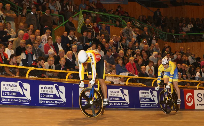 Two cyclists competing on an indoor velodrome track in Bordeaux, riding on the banked surface in front of a large crowd. French Cycling Federation signage visible.