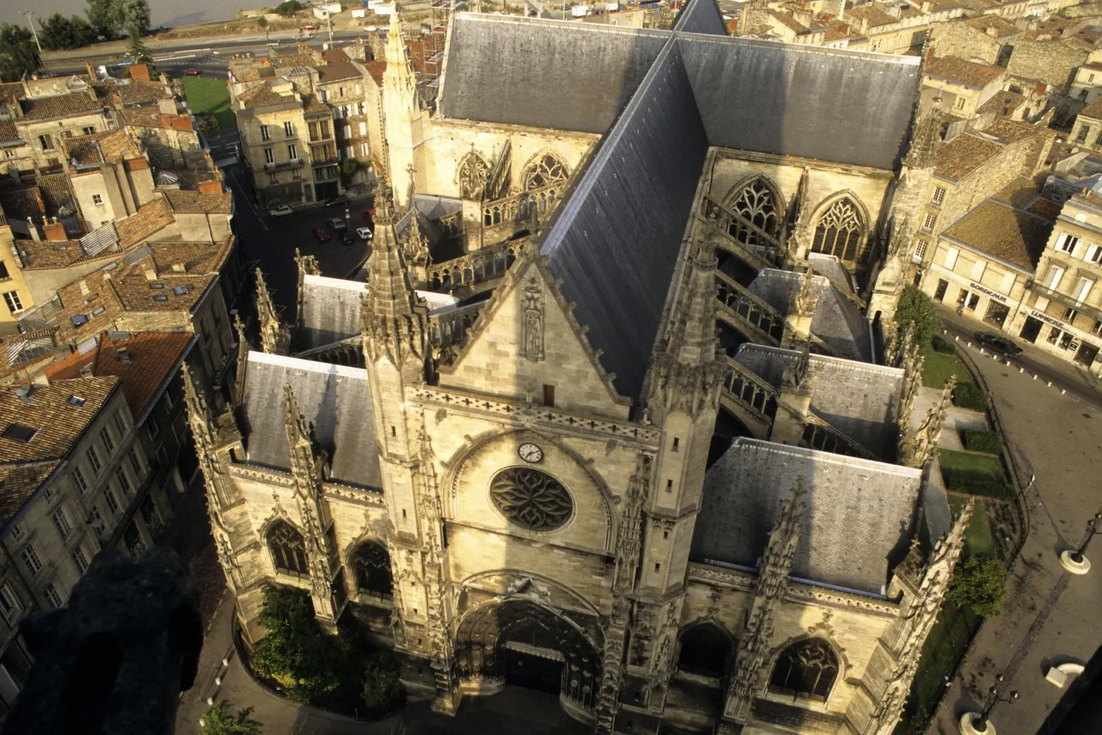 Aerial view of the Basilica of Saint-Michel in Bordeaux displaying Gothic architecture with slate roofs, stained glass windows, and a bell tower, surrounded by urban buildings.