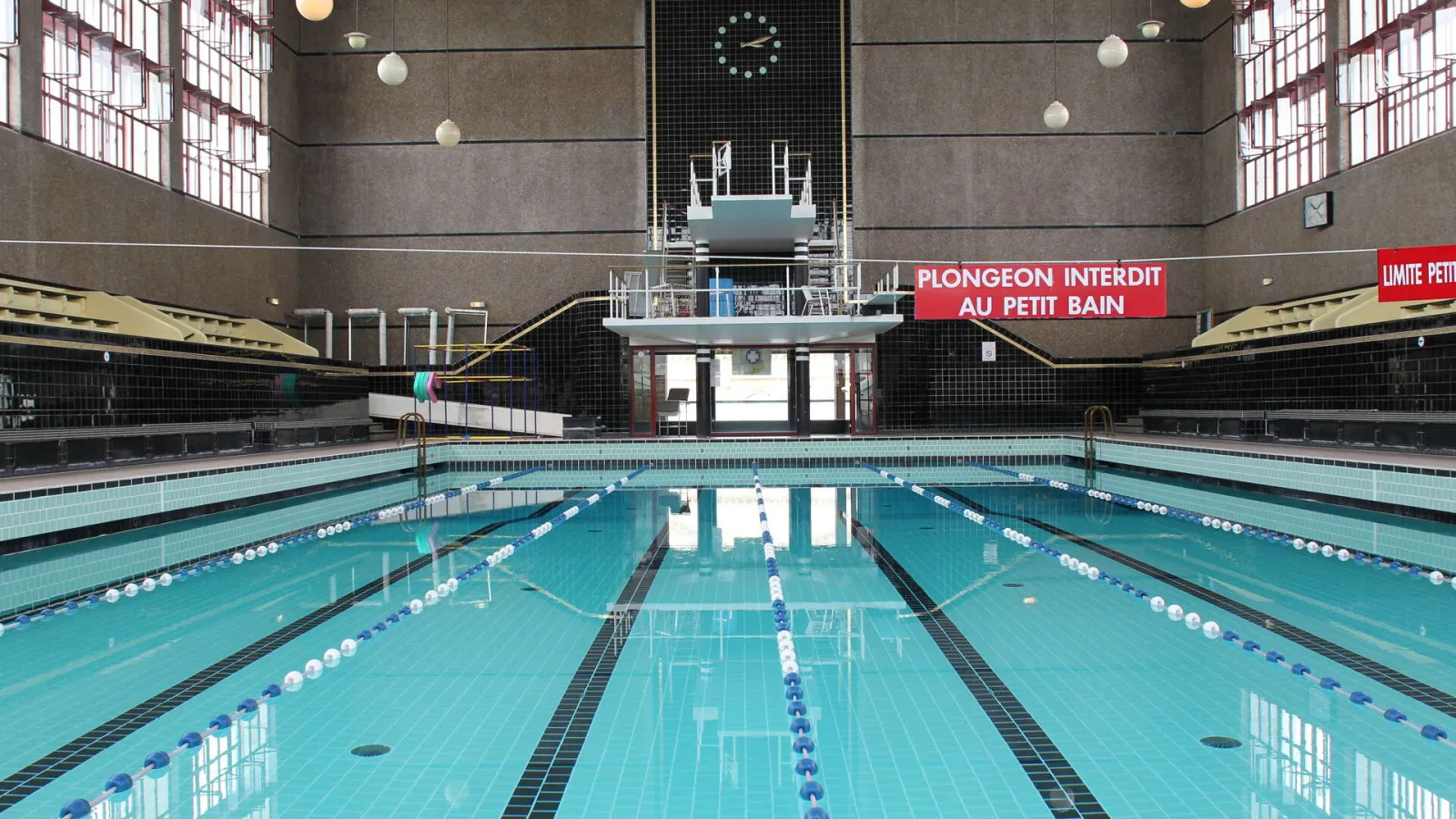Indoor Olympic swimming pool with three lanes marked by lane dividers in a large hall with gray and black walls and windows, at the Judaïque Jean Boiteux municipal pool in Bordeaux.