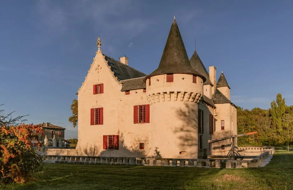 Château Olivier, a medieval-style castle with pale pink walls, pointed turrets, and red windows, surrounded by green parkland near Bordeaux.