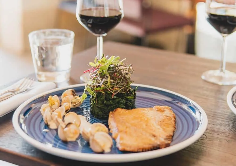 Gourmet plate featuring golden fish, shrimp, and artistic green salad presentation on blue and white ceramic, paired with red wine glasses in an upscale dining setting.
