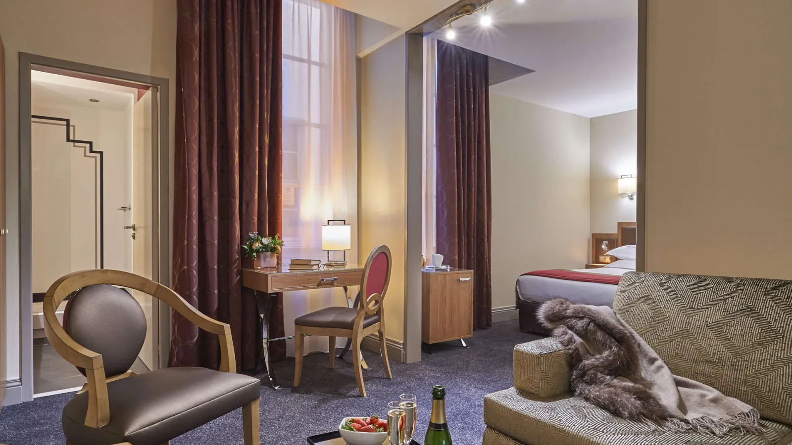 Luxury suite at Best Western Premier Hôtel Bayonne Etche Ona in Bordeaux, featuring a king-size bed, lounge area with desk, elegant armchair, burgundy curtains, and natural wood furniture