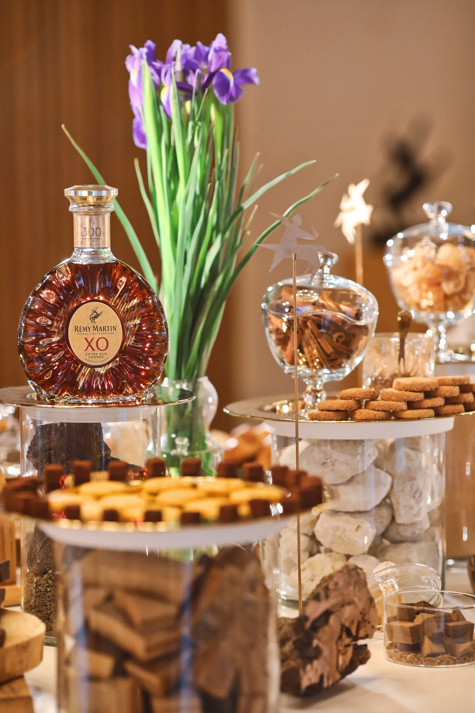 An elegant display of a Rémy Martin XO cognac bottle surrounded by pastries, crackers, and candies in glass bowls, with a vase of purple irises in the background at Rémy Martin House in Bordeaux.