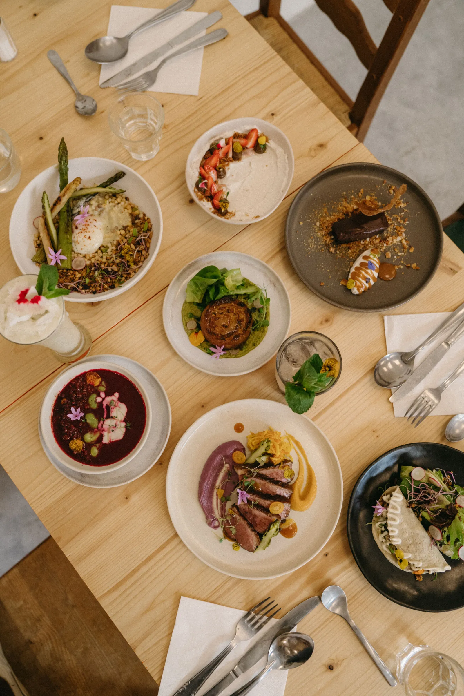 Restaurant table setting featuring gourmet dishes including asparagus risotto, beet soup, duck confit, seared seafood in a pan, and refined culinary creations garnished with edible flowers and fresh herbs