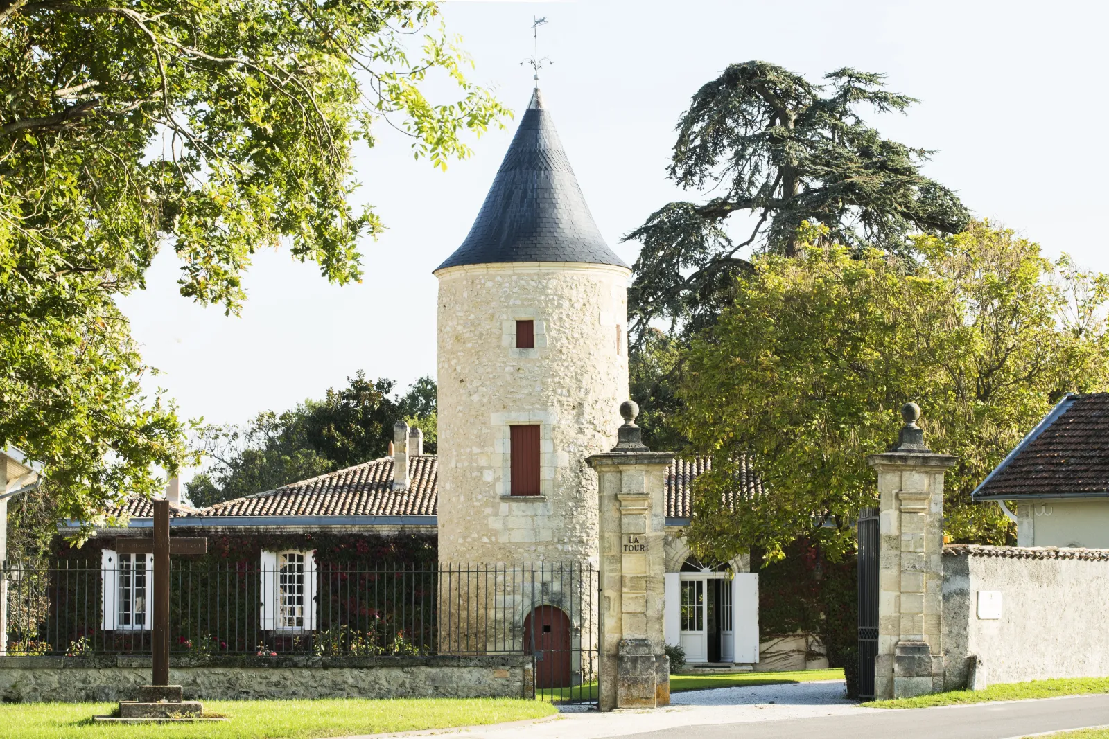 Latour-Martillac Castle in Bordeaux featuring a distinctive white stone tower with conical black roof, wrought iron gate entrance, and surrounding heritage buildings nestled in lush greenery.