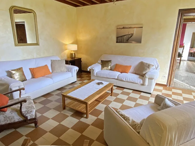 Elegant living room at Château La Gontrie in Bordeaux featuring two white sofas, beige armchairs, wooden coffee table, checkered beige and white tile flooring, pale yellow walls, gold mirror and classic furnishings.