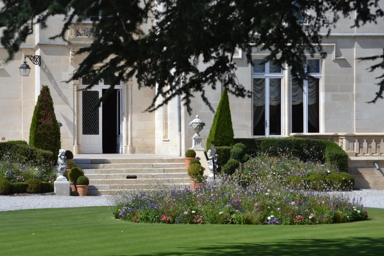 Classical facade of Château Pape Clément in Bordeaux with formal French garden, lavender and rose flower beds, topiary bushes, stone stairs and architectural ornaments.