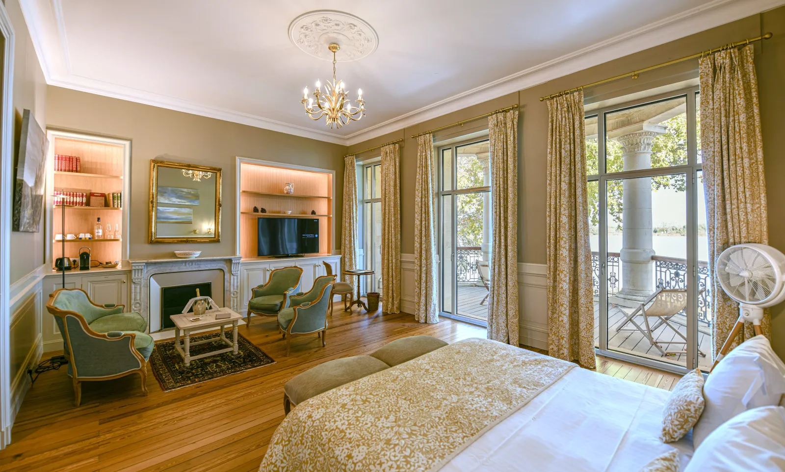 Elegant suite at Château Grattequina in Bordeaux featuring a king-size bed, private sitting area, fireplace, Louis XV furniture, gold curtains and a terrace overlooking the vineyards.