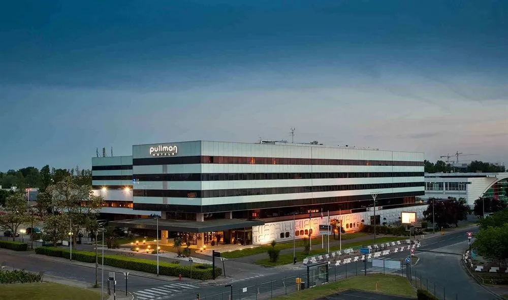 Pullman Bordeaux Lac hotel, modern building with striped white and black façades, lit up in the evening with landscaped forecourt and road access