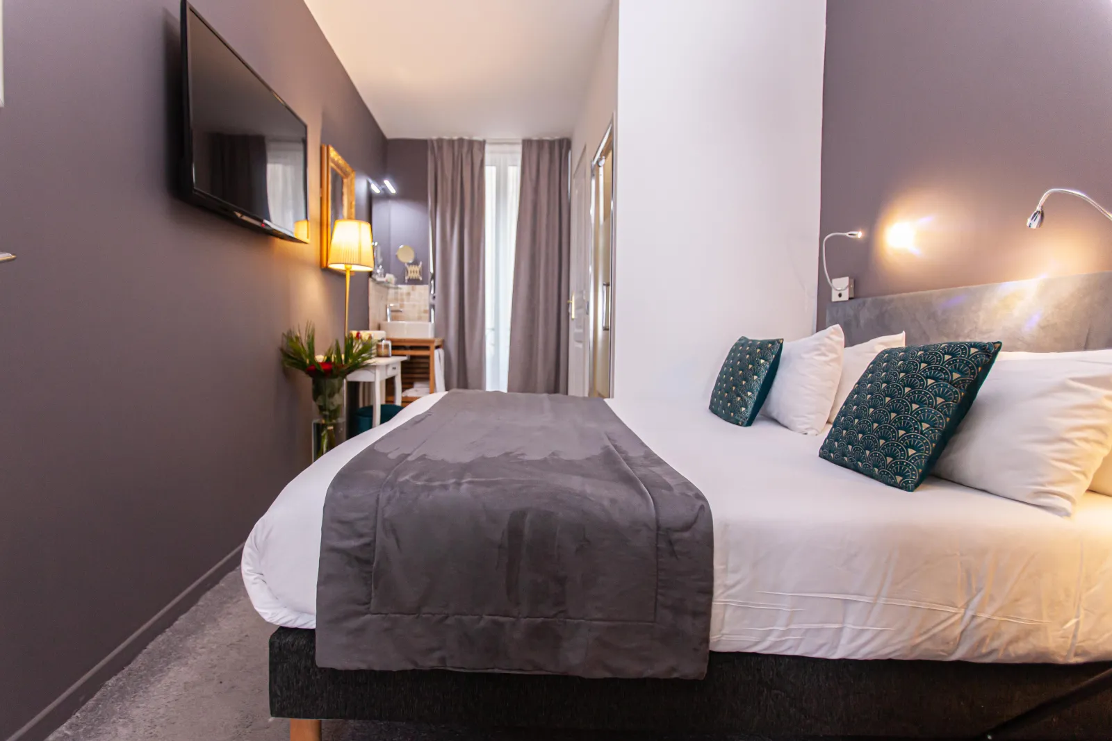 Modern hotel bedroom featuring a queen-size bed, mauve-grey walls, warm lighting, turquoise patterned pillows, and glass doors opening onto a balcony at Hotel Churchill in Bordeaux.