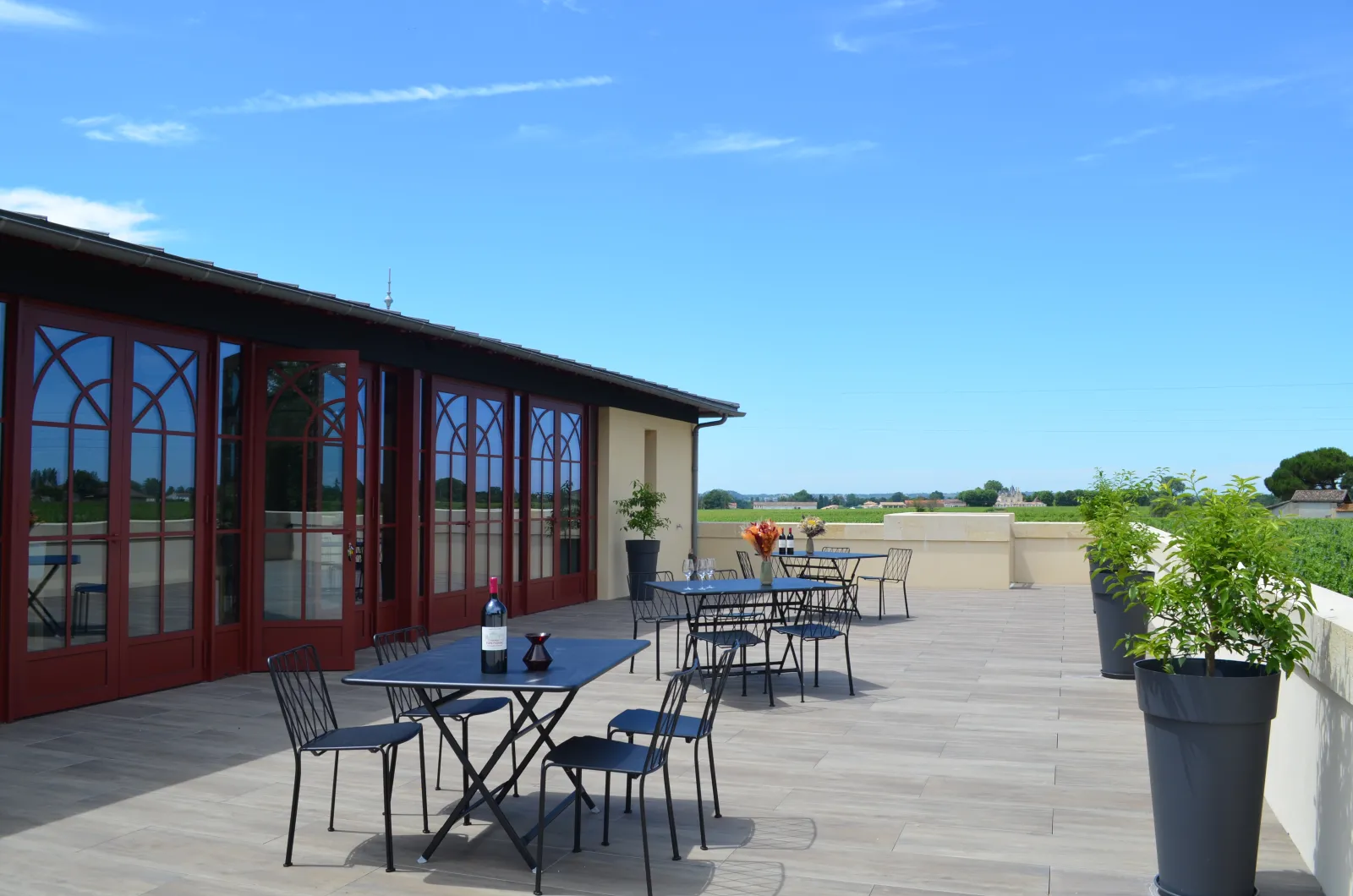 Terrace of Château Yon-Figeac in Bordeaux featuring blue tables and chairs, red-doored building overlooking the local vineyards
