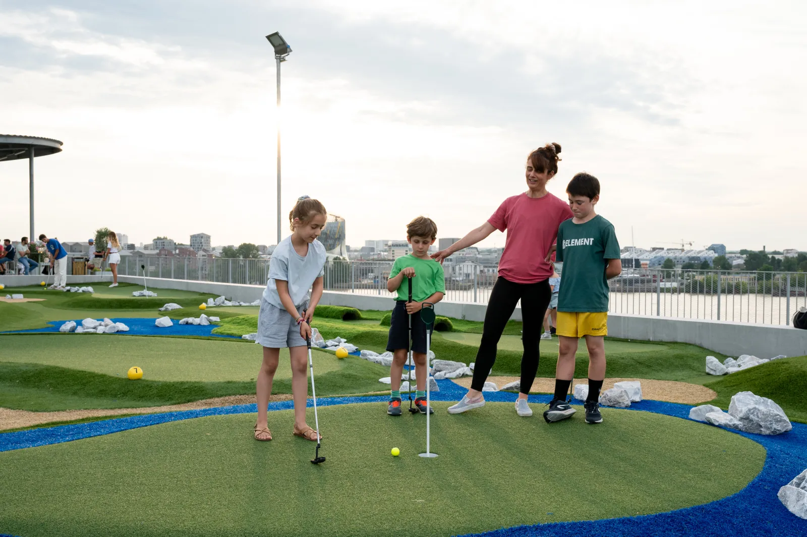 A woman and three children playing mini-golf on an elevated course overlooking Bordeaux at the UCPA Sport Station rooftop.