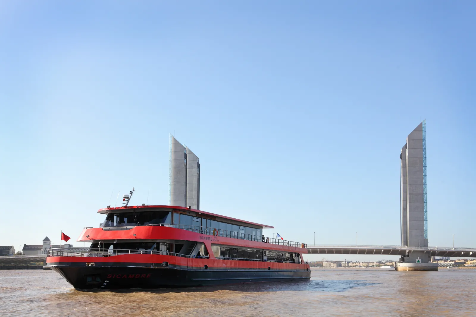 Red and blue cruise boat sailing on the Garonne river in Bordeaux, with the Water Mirror towers visible in the background under clear skies.