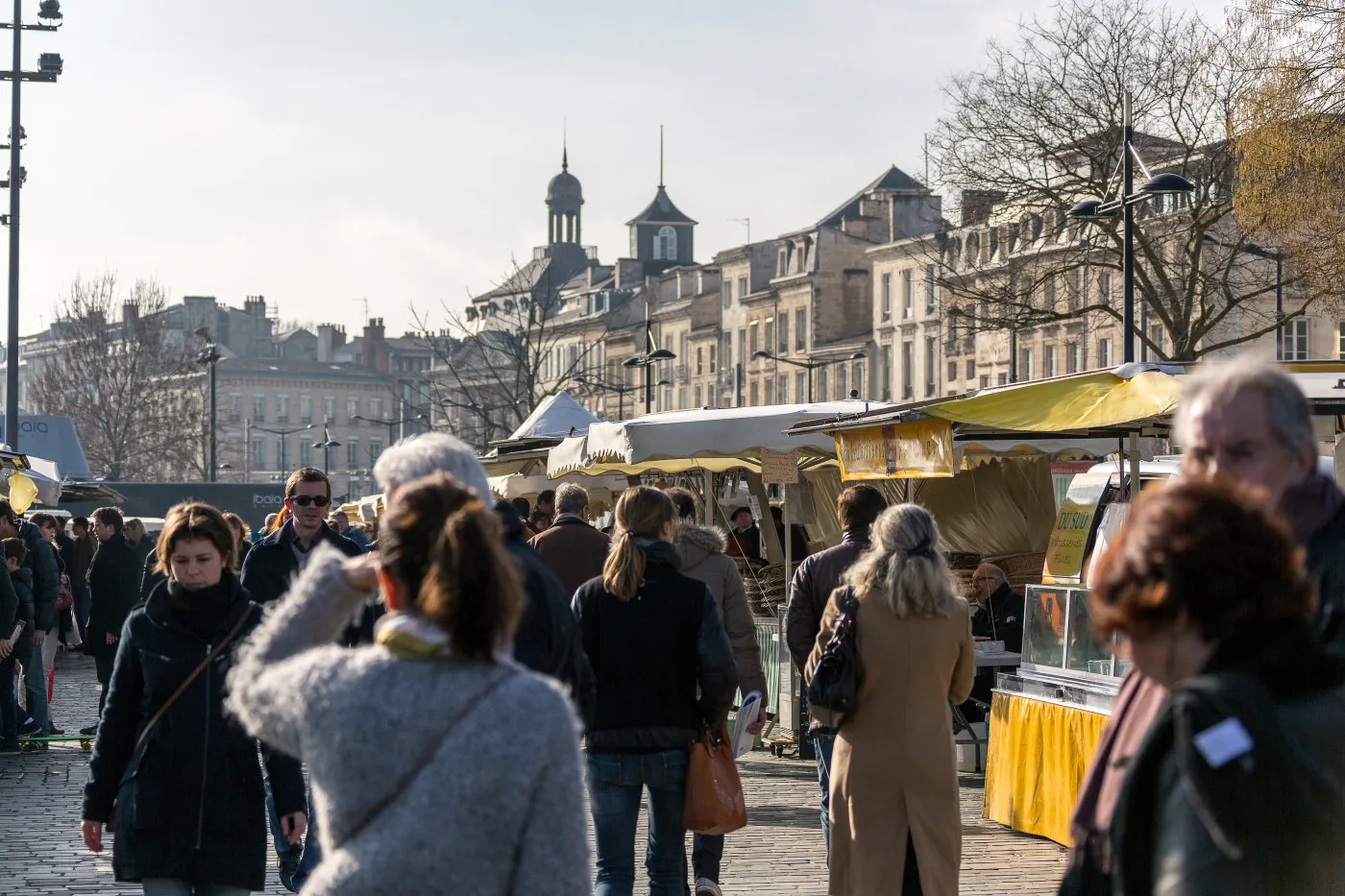 Bustling market at Quai des Chartrons in Bordeaux with visitors browsing yellow market stalls, Haussmann-style buildings and churches in the background.