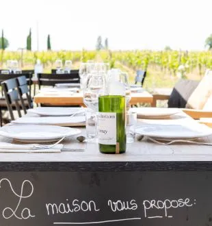 Outdoor dining table overlooking Bordeaux vineyards with white wine bottle, glasses, place settings, and blackboard sign reading 'La maison nous propose'