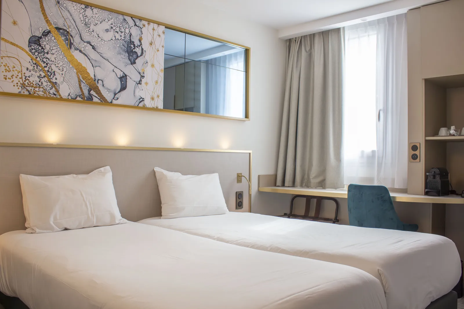 Modern hotel room featuring a double bed with white linens, gray upholstered headboard with built-in lighting, gold-framed mirror above, desk with blue chair, window with beige curtains, and abstract gold and gray wall art.