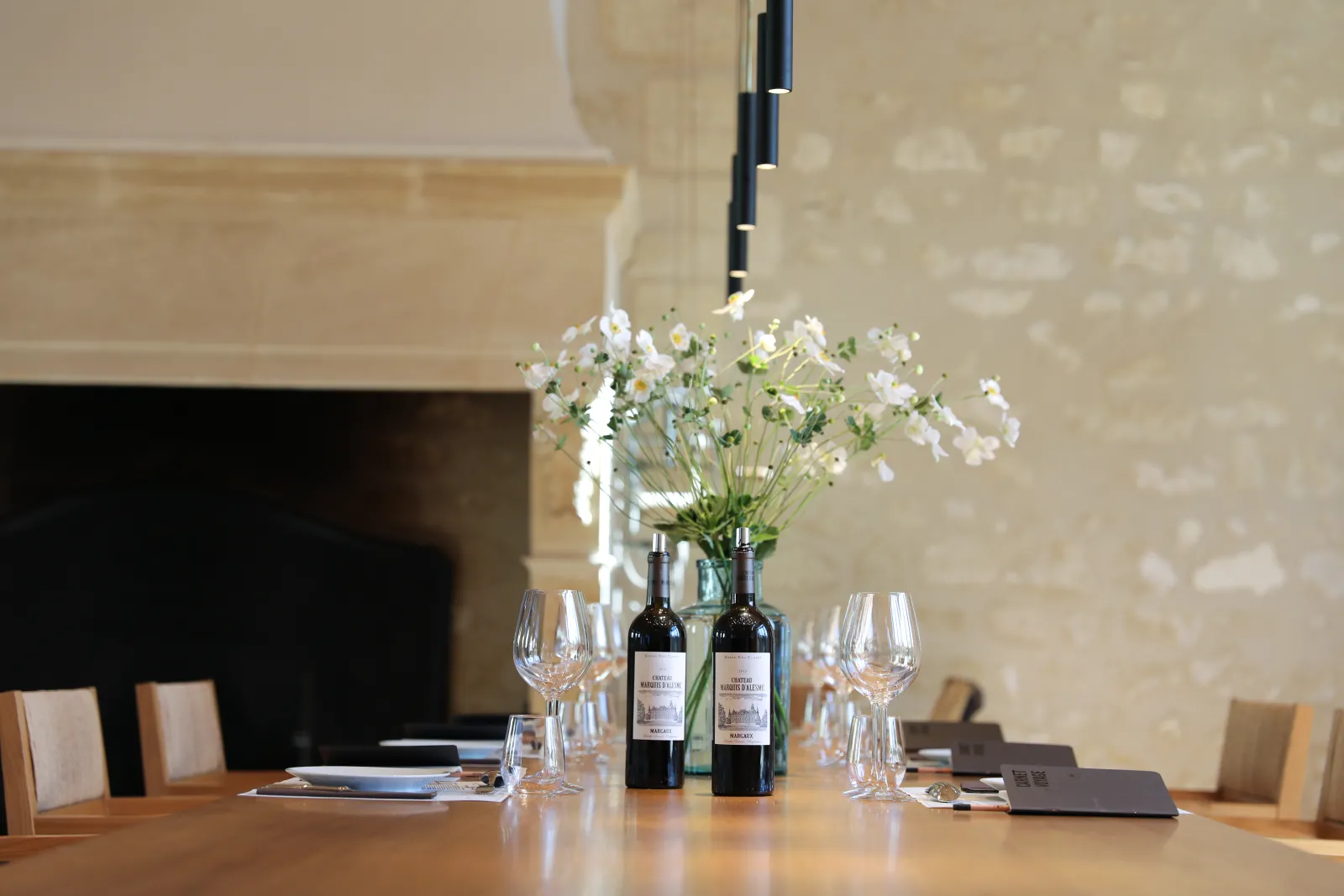 Elegant dining table with wine glasses, Bordeaux wine bottles, white flowers in a vase, and place settings in an upscale restaurant.