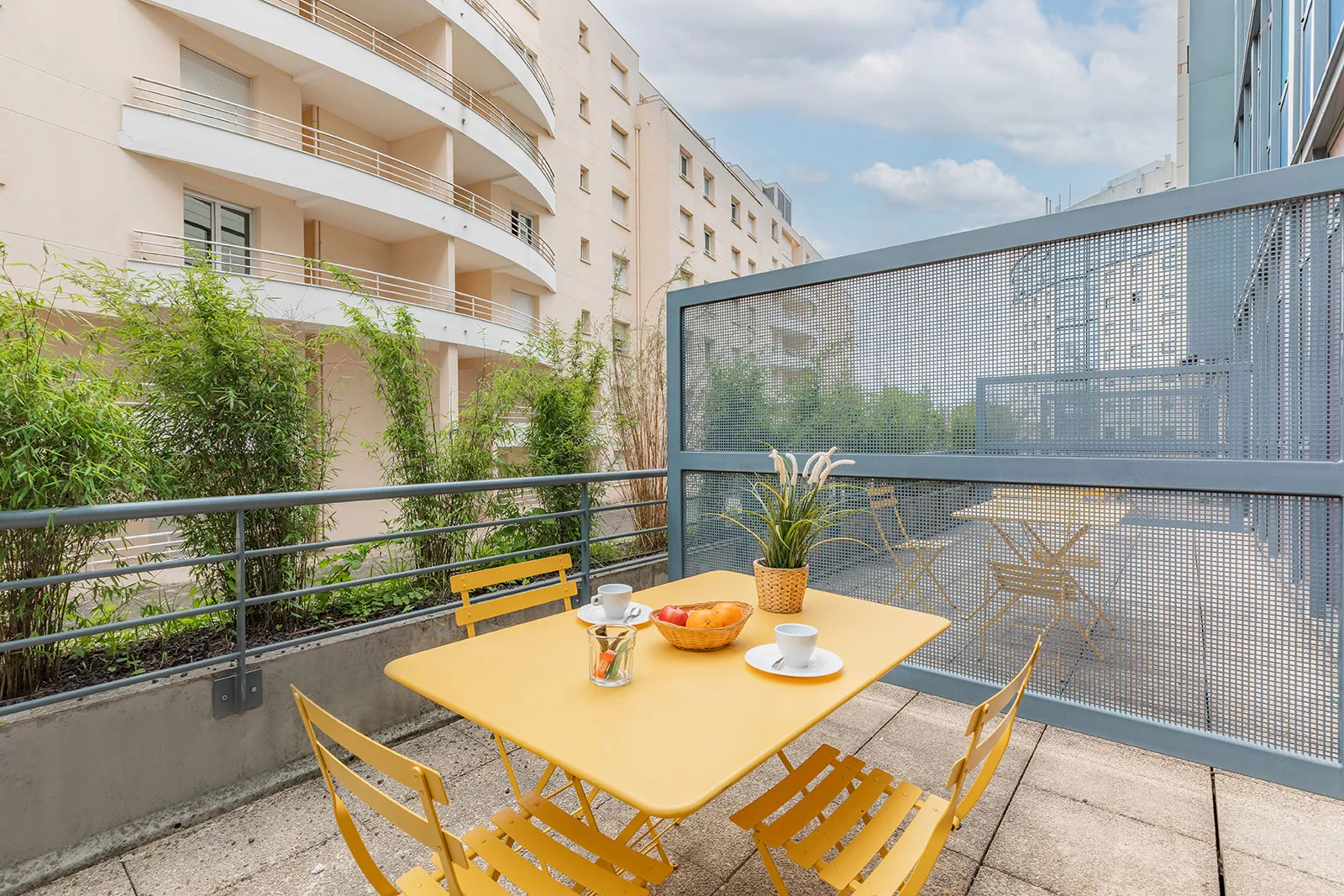 Sunny terrace of an apartment in Bordeaux with yellow table and chairs, overlooking residential buildings and surrounding greenery