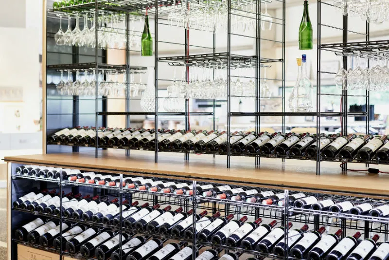 Modern wine cellar with metal shelving displaying red and white wine bottles, hanging glasses, and wooden counter in the foreground at Château Prieuré-Lichine in Bordeaux.