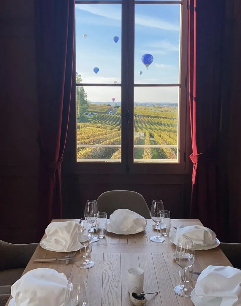 Elegant dining room table set with white napkins and glasses overlooking Bordeaux vineyards with hot air balloons floating in the blue sky