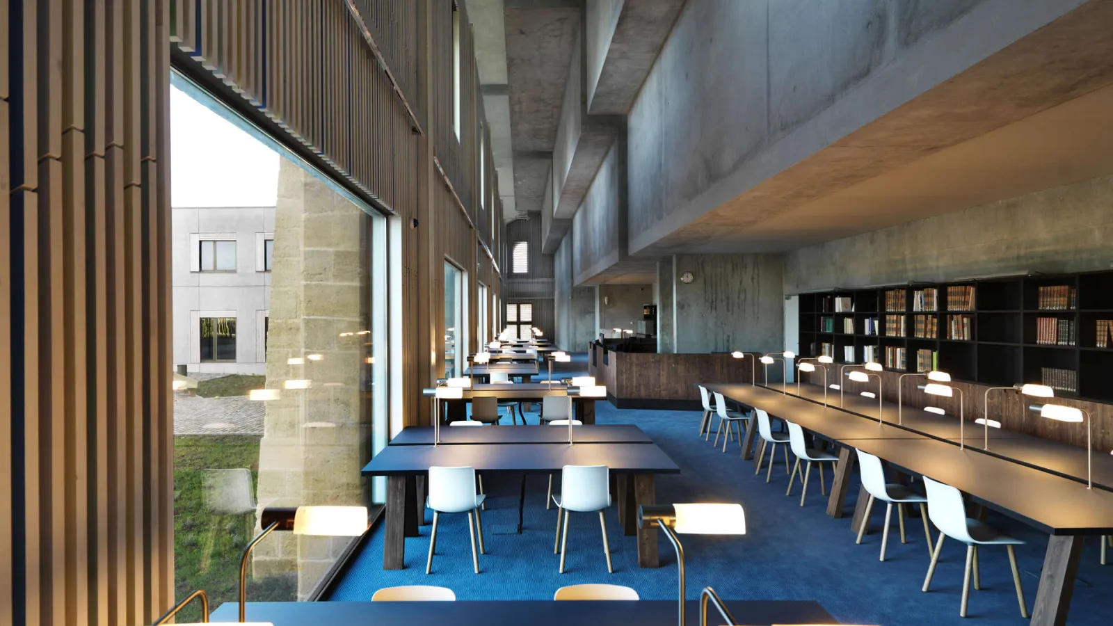 Modern library interior featuring wooden work tables, white and blue chairs, bookshelves, blue carpet flooring, large glass windows overlooking a patio, concrete ceiling and contemporary architectural design.