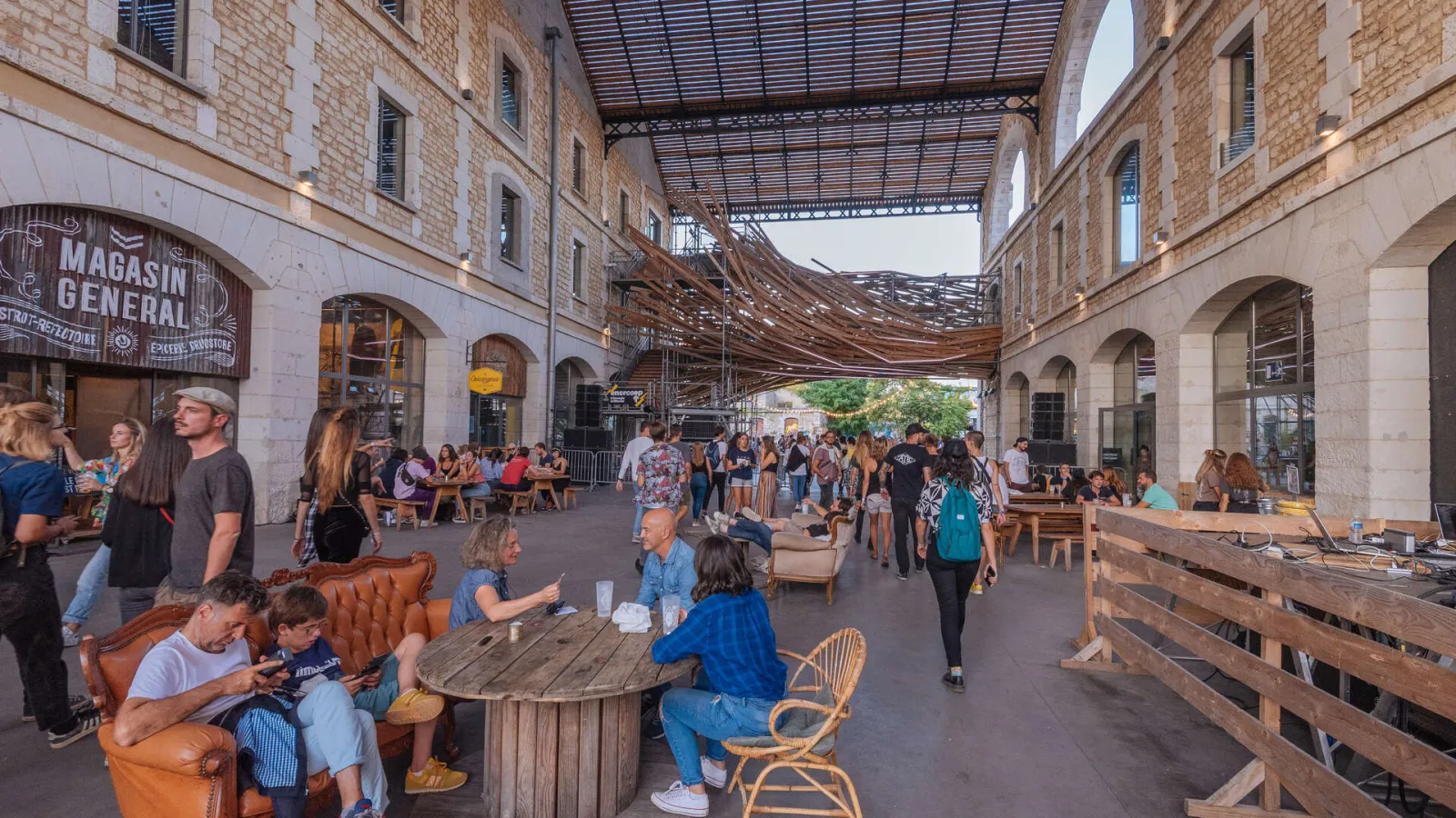 Interior view of Darwin, Bordeaux's covered public space featuring restaurants, bars and shops. The pedestrian area hosts visitors seated at tables and chairs, with brick architecture, arcades, and an open metal roof.