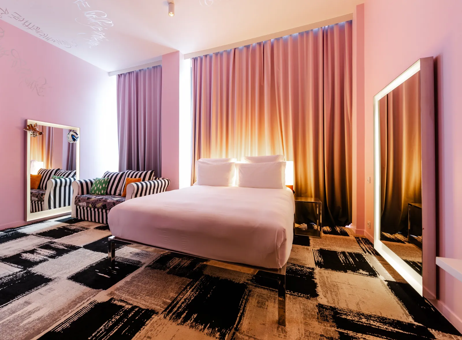 Modern bedroom at Mama Shelter Bordeaux featuring a white bed, soft pink walls, gradient orange and gold curtains, illuminated mirrors, striped black and white armchair, and geometric black and beige carpet