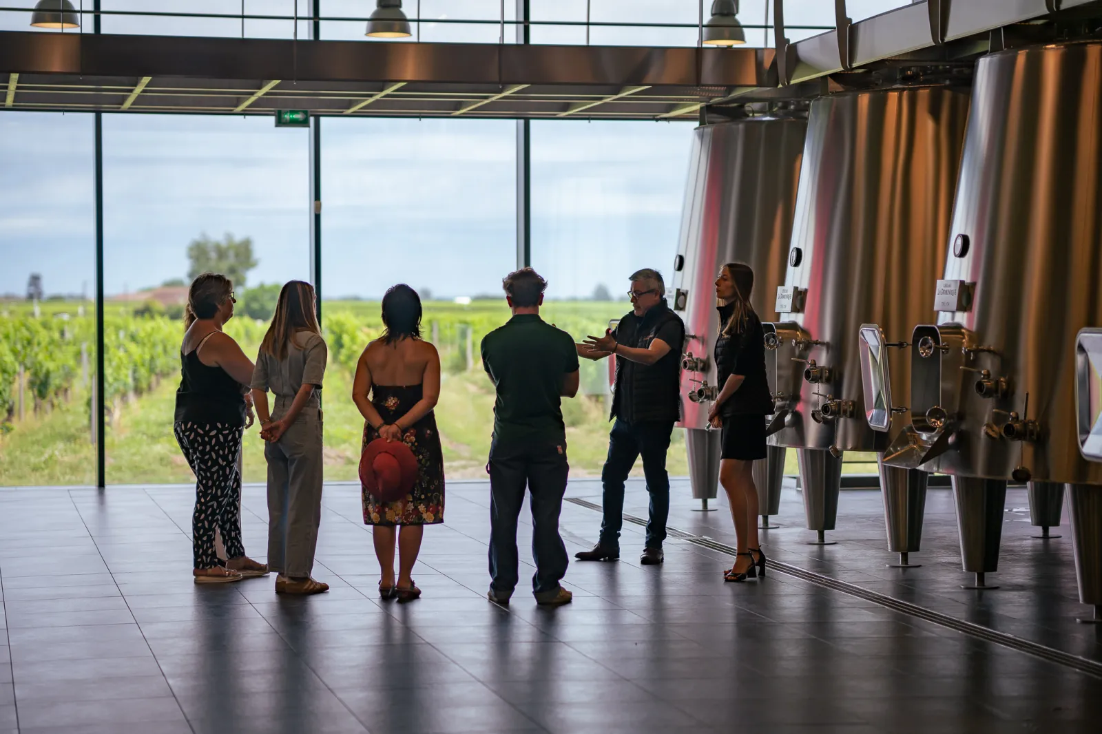 Group of six visitors on a guided tour in a modern winery facility with stainless steel tanks, overlooking Bordeaux vineyards through large panoramic windows.