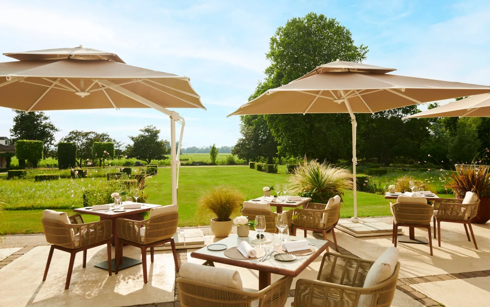 Elegant outdoor dining terrace at Grand Barrail restaurant in Bordeaux featuring set tables, rattan chairs, beige umbrellas and views of lush green gardens