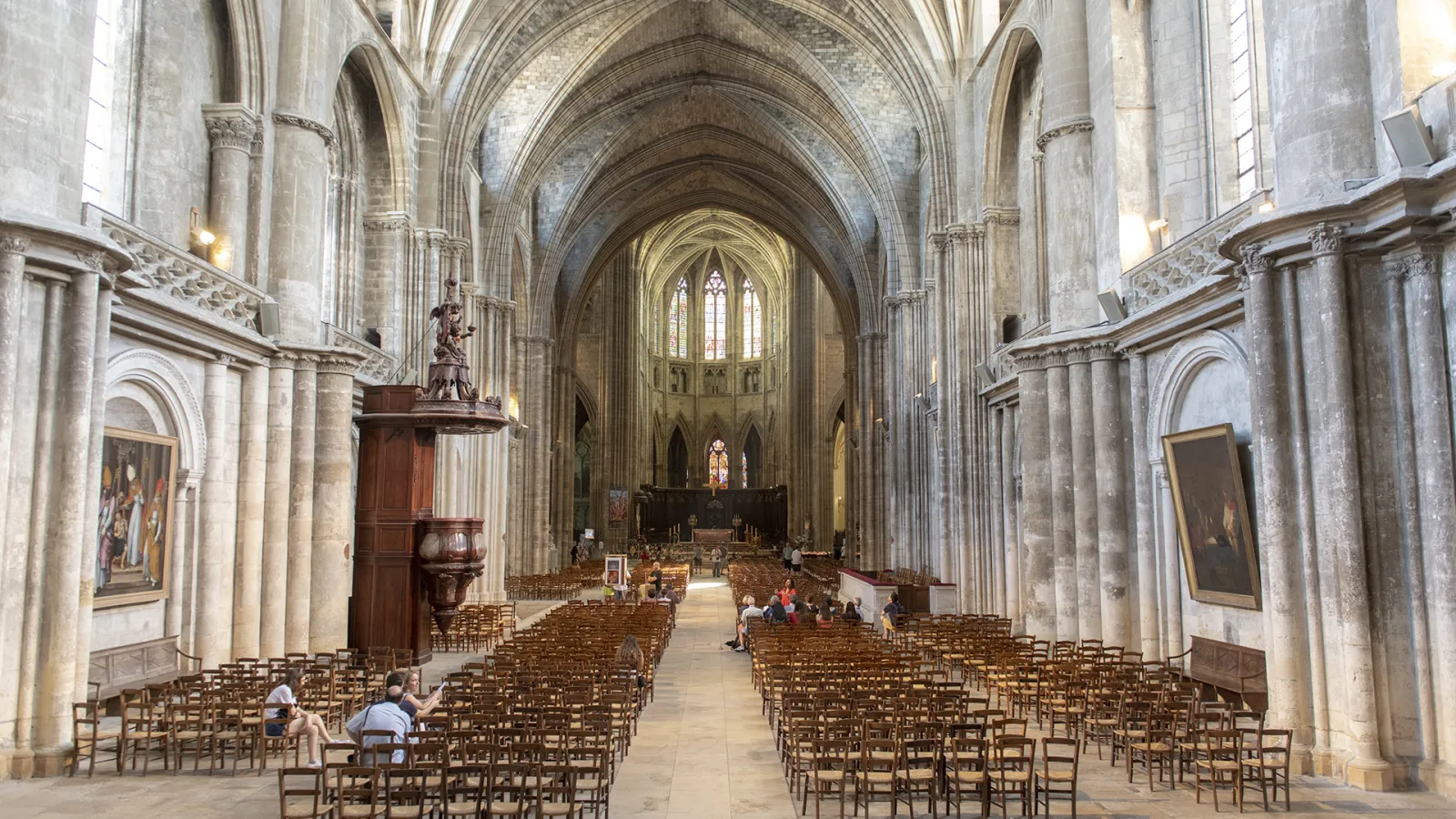 Interior of Bordeaux Cathedral of Saint-André featuring pointed Gothic vaults, massive columns, wooden chairs arranged in the nave, and the altar illuminated by stained glass windows.