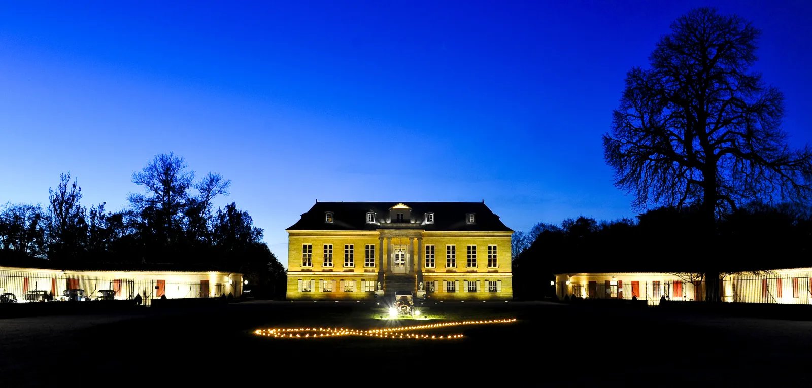 Château La Louvière in Bordeaux lit up at night, classical yellow façade with black mansard roof, flanked by side buildings and bare trees against a twilight blue sky.