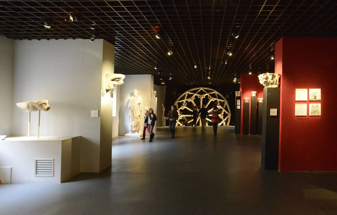 Interior of the Musée d'Aquitaine in Bordeaux featuring a spacious exhibition gallery with white and red walls, ancient sculptures on display, a large circular golden art installation in the center, and visitors exploring the collections.