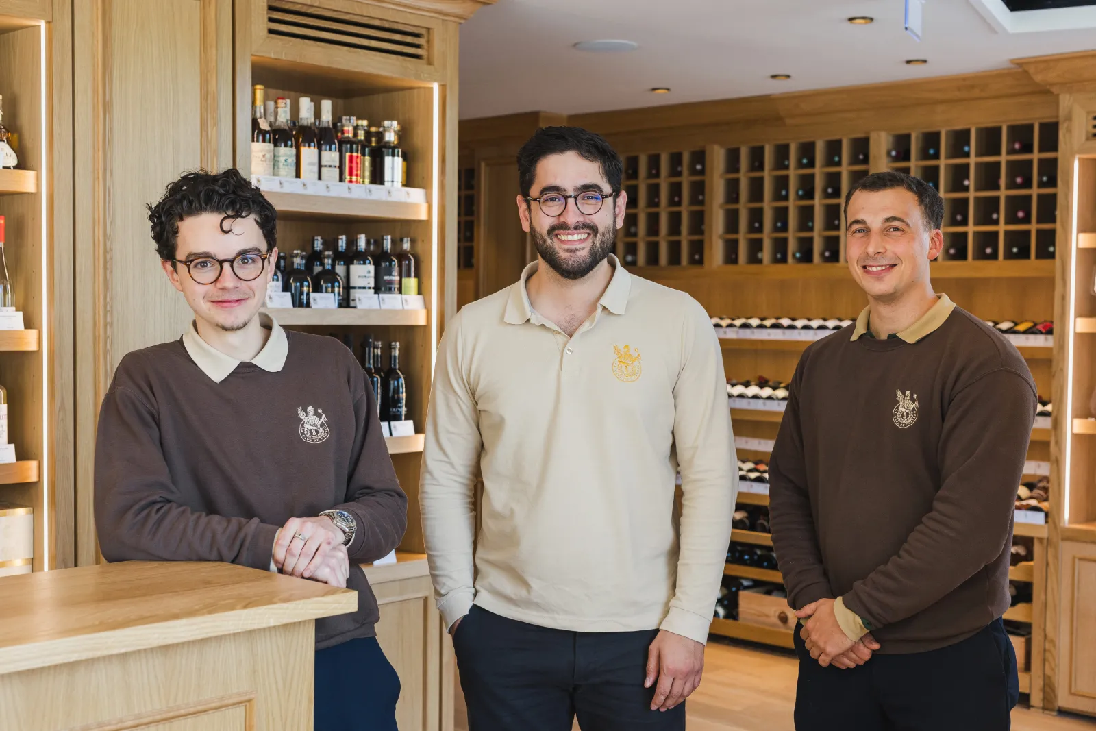 Three smiling men pose in a Bordeaux wine cellar with wooden shelves of bottles in the background. They wear sweaters with a distinctive logo.