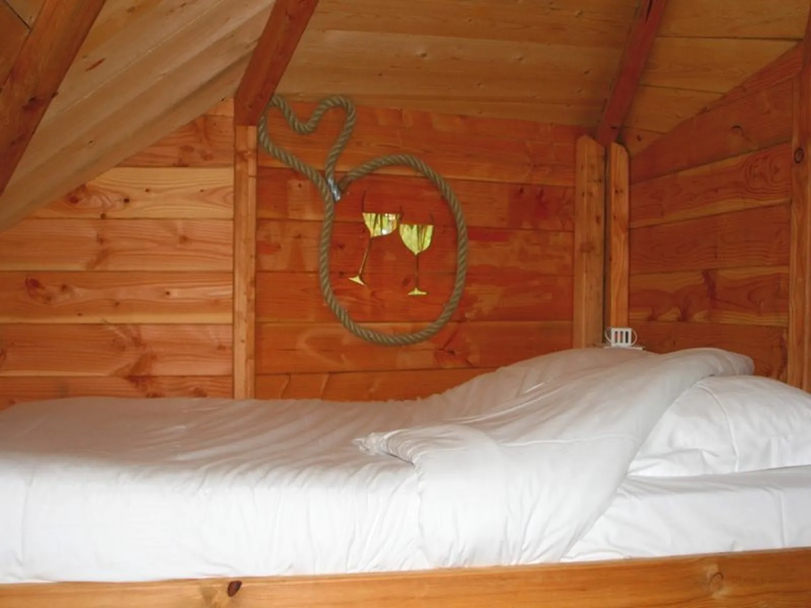 Interior of a wooden cabin featuring a bed with white linens and wall decoration showing a stylized face made from rope and golden leaves
