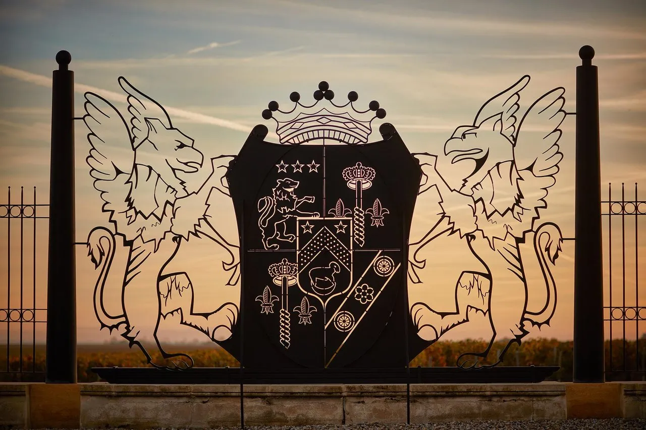 Coat of arms of Château Pichon Baron in Bordeaux, black silhouette featuring two winged dragons, crown and heraldic shield against a sunset sky backdrop