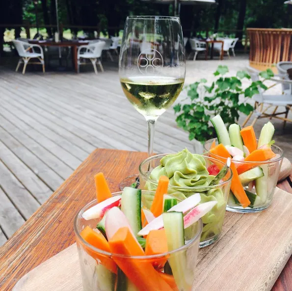 Glass of white wine on a terrace with fresh vegetable sticks (carrots, cucumber, radishes) in glasses, wooden table, view over Château Léognan park in Bordeaux